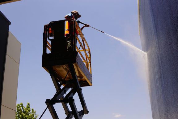 Un ouvrier nettoie une surface verticale depuis un élévateur mobile sous un ciel bleu.